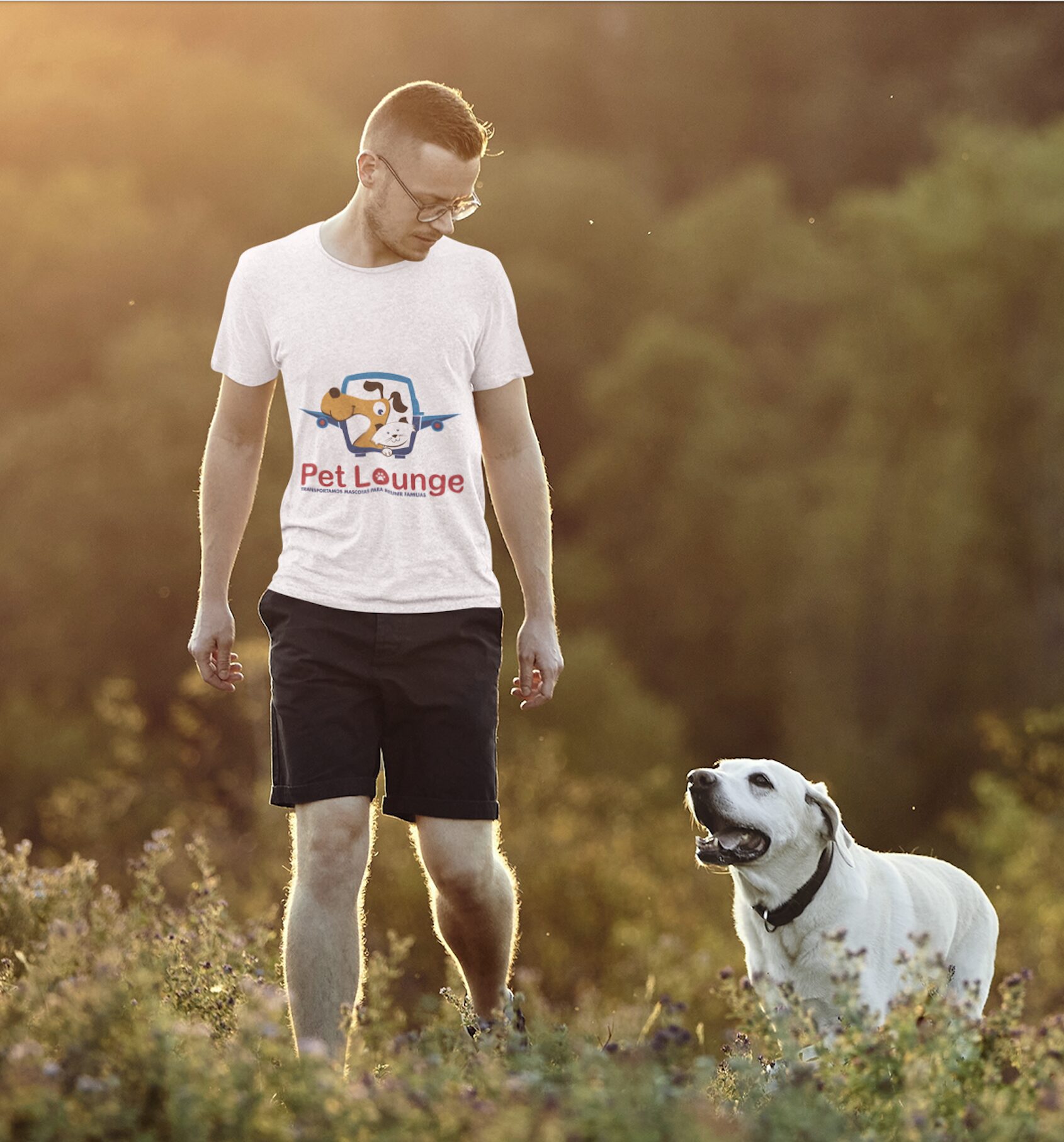 A man in a "Pet Lounge" T-shirt walking in a field with his Labrador retriever at sunset.